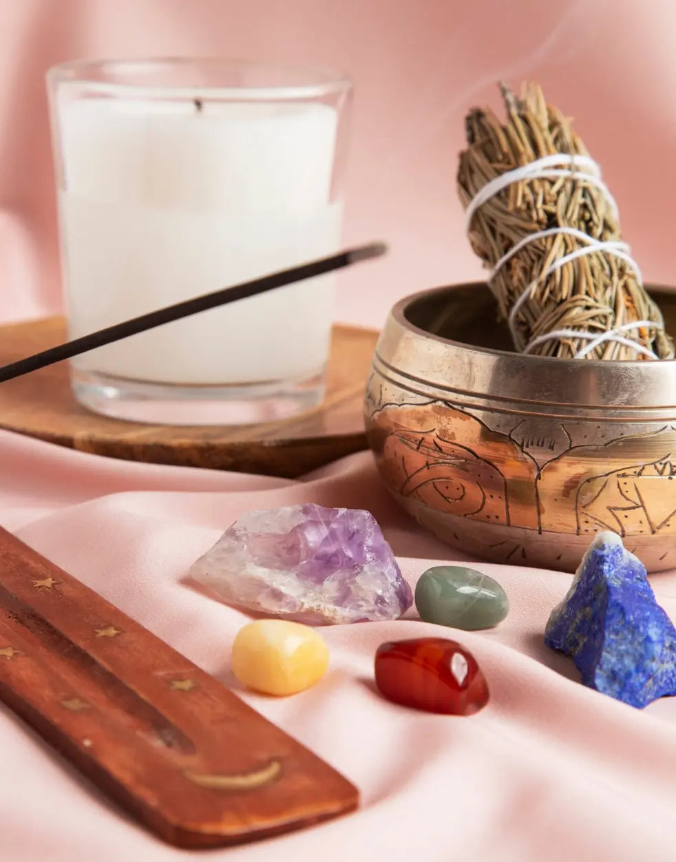 White candle, sage bundle, singing bowl and healing crystals on pink fabric
